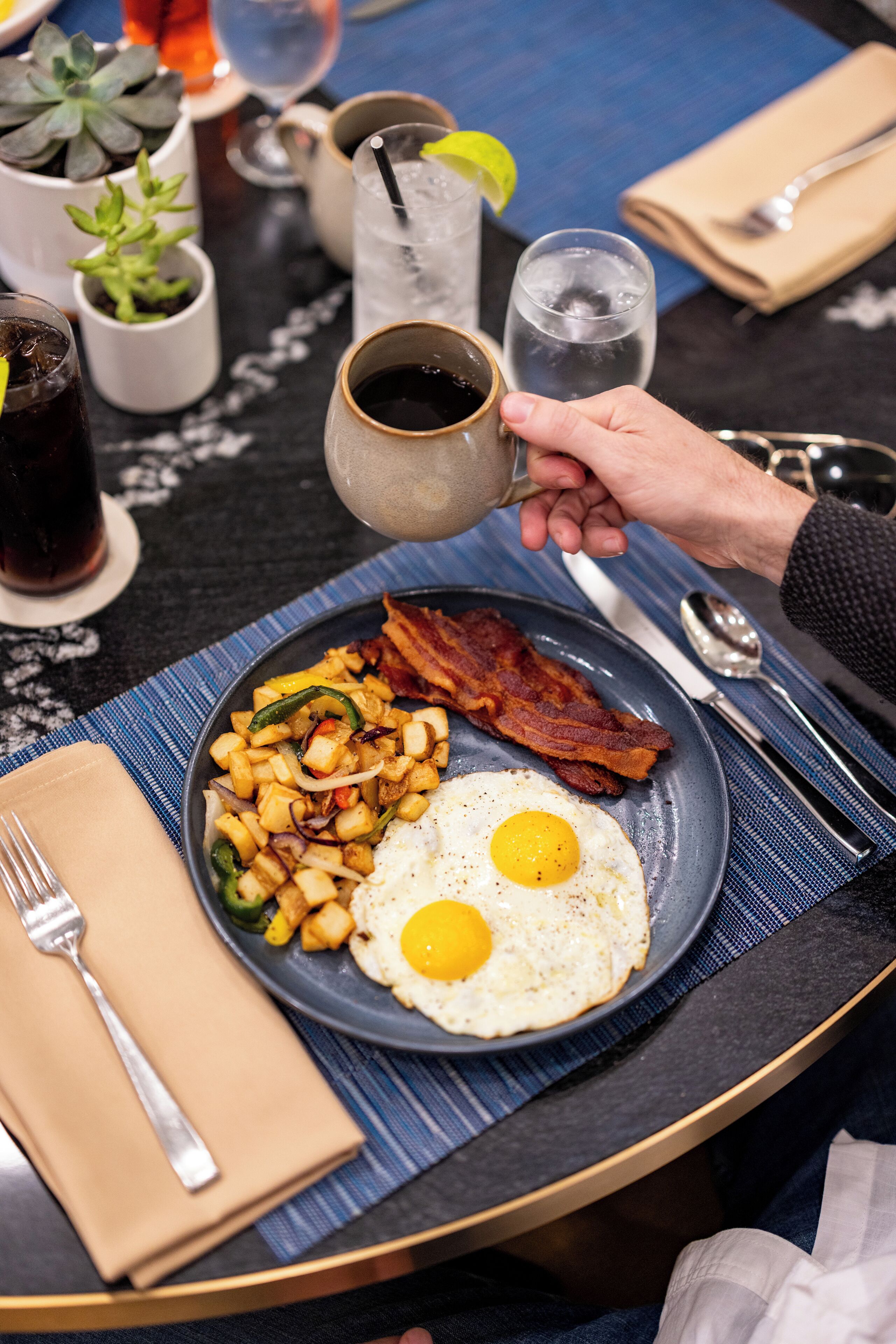 Un plato de desayuno con huevos, tocino y papas, una taza y un vaso de agua helada sobre una mesa.
