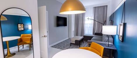 A modern hotel room with a large mirror, a round dining table, a sofa, and a TV mounted on the wall.