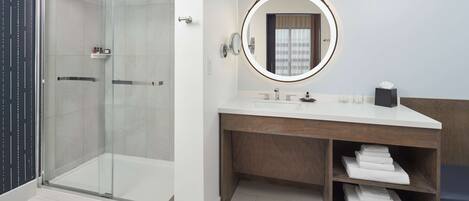 A modern bathroom with a glass-enclosed shower, a vanity with a wooden cabinet, and a round mirror with a light.