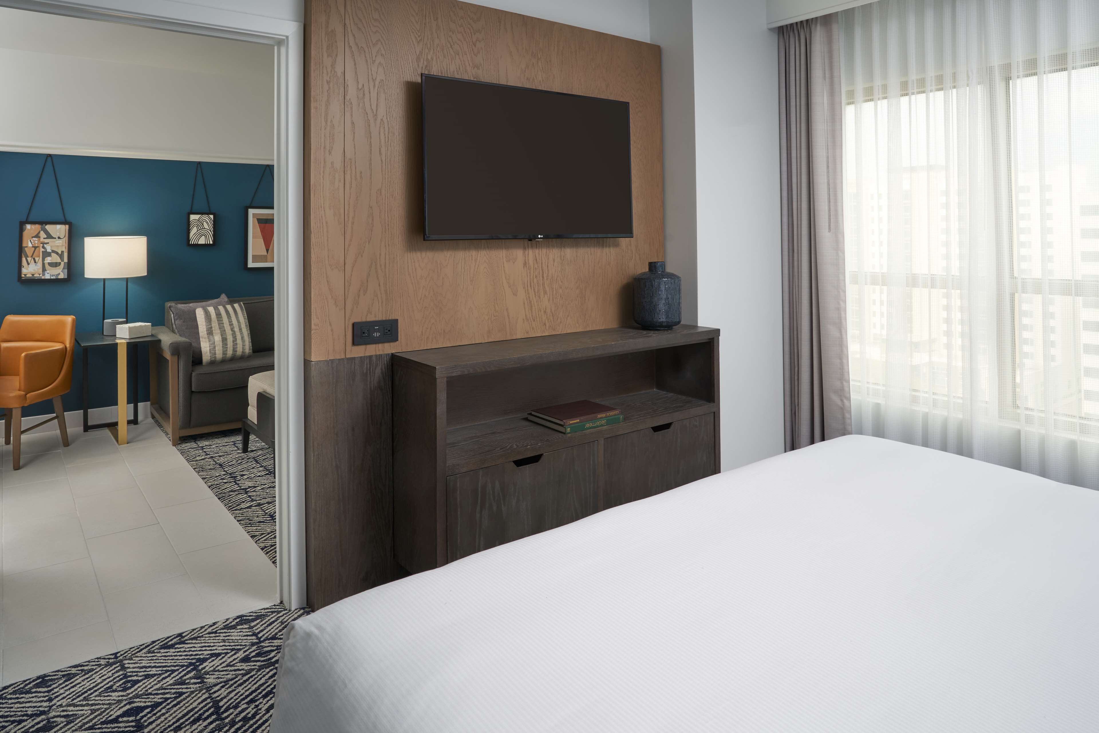 Une chambre d’hôtel dotée d’un grand lit, d’un téléviseur fixé sur un panneau en bois, d’un coin salon avec un canapé et un fauteuil, et offrant une vue sur la ville à travers des rideaux transparents.