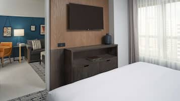 Een hotelkamer met een groot bed, een televisie bevestigd aan een houten paneel, een zithoek met een bank en fauteuil, en uitzicht op de stad door dunne gordijnen.