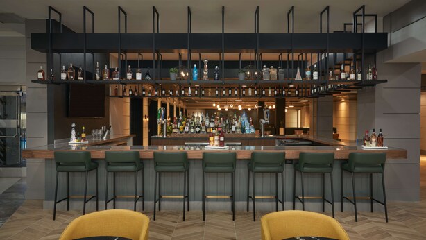 Un área de bar moderna con múltiples estantes llenos de botellas de licores, una barra central y asientos con mesas y sillas.