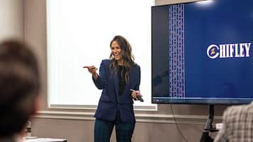 A conference room with a presentation screen displaying the Chifley logo, a speaker addressing an audience, and attendees seated at tables with laptops.