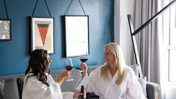 Two people in white robes sitting on a grey sofa in a room with blue walls, a wooden tray with snacks, and a bottle of wine.