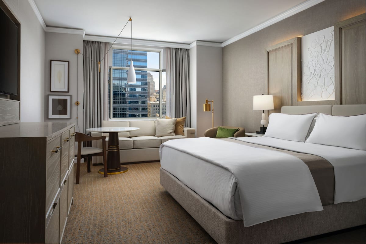 A hotel room with a large bed, a desk, a chair, a small table, a window with curtains, and a city view.