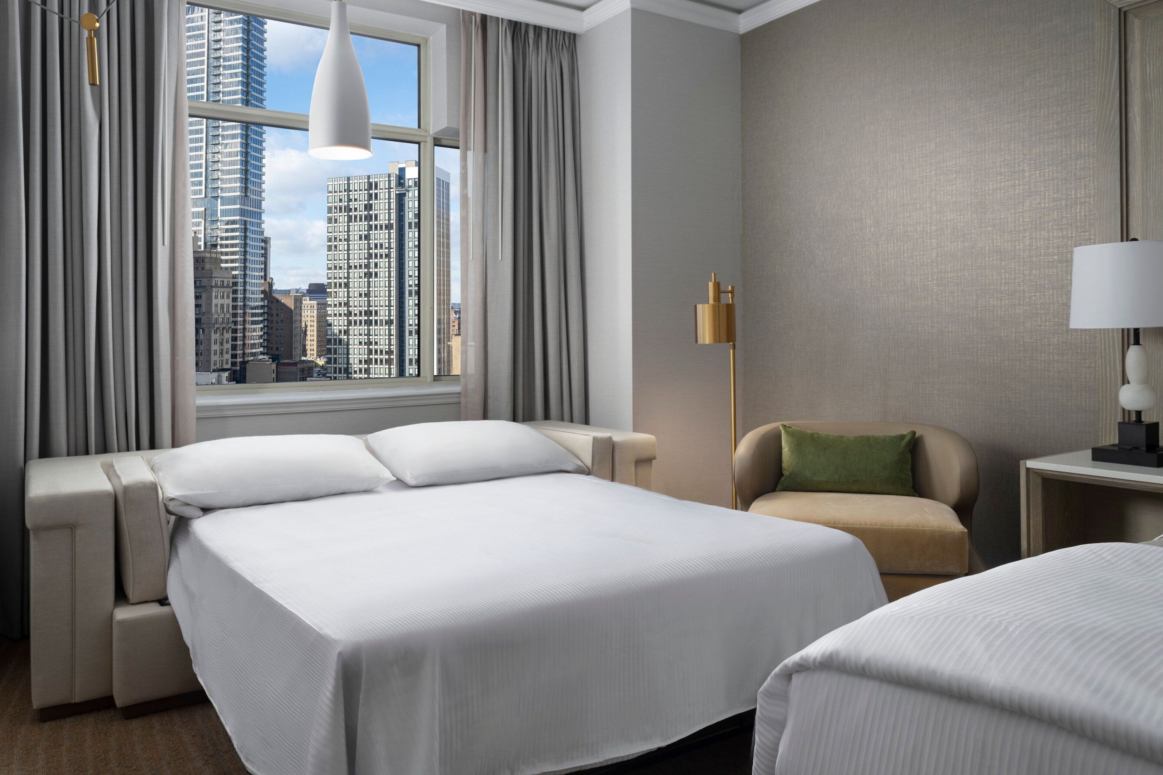 Une chambre d’hôtel avec un grand lit, une chaise, une lampe et une vue sur le paysage urbain par la fenêtre.