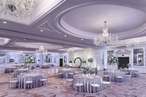 A spacious banquet hall with round tables, clear chairs, and elegant chandeliers.