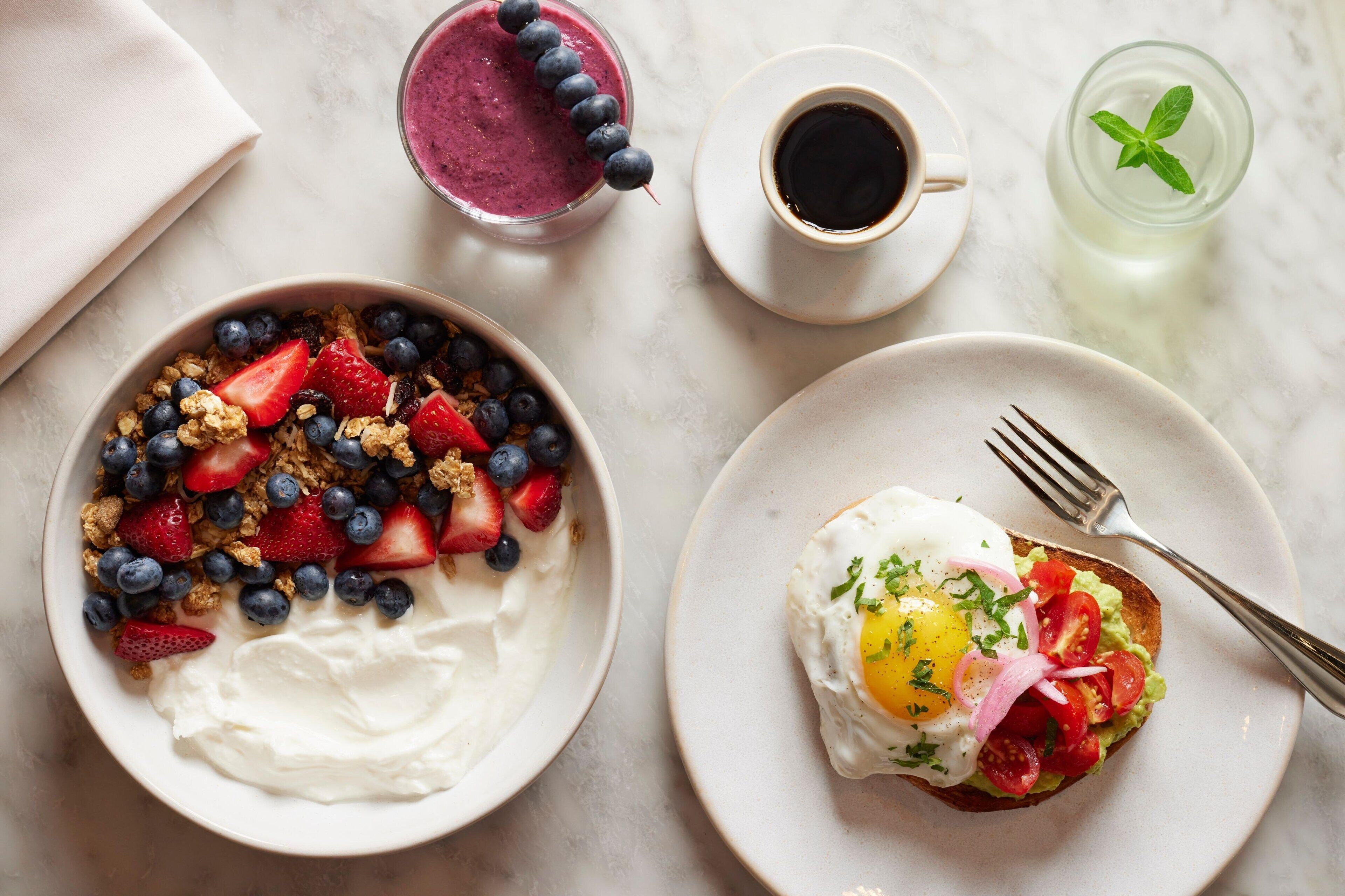A breakfast setting with a bowl of yogurt topped with strawberries and blueberries, a plate with an egg on toast, a cup of coffee, and a glass of iced tea.
