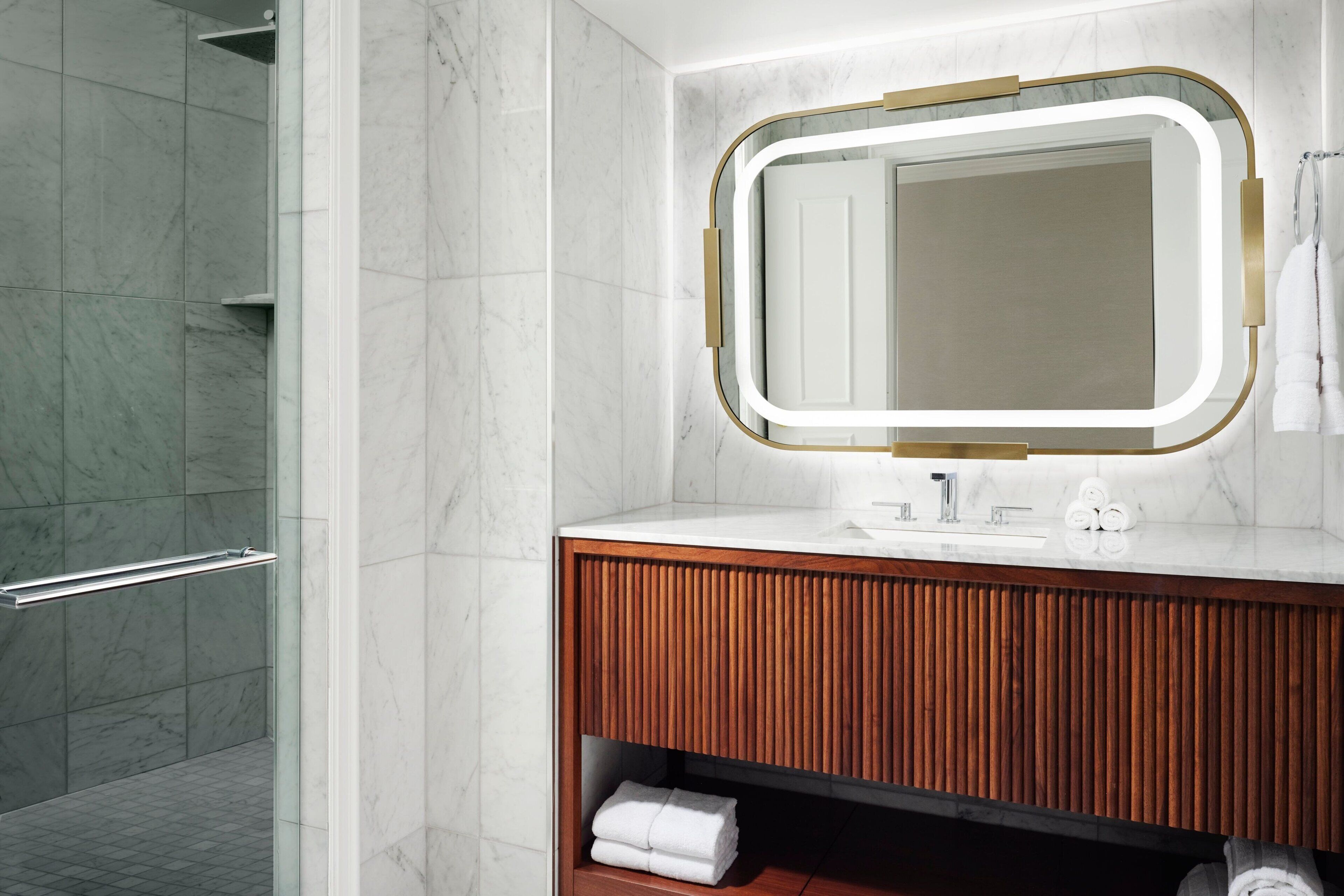 Een badkamer met een houten wastafelmeubel, een spiegel met een licht frame en een douche met een glazen deur.