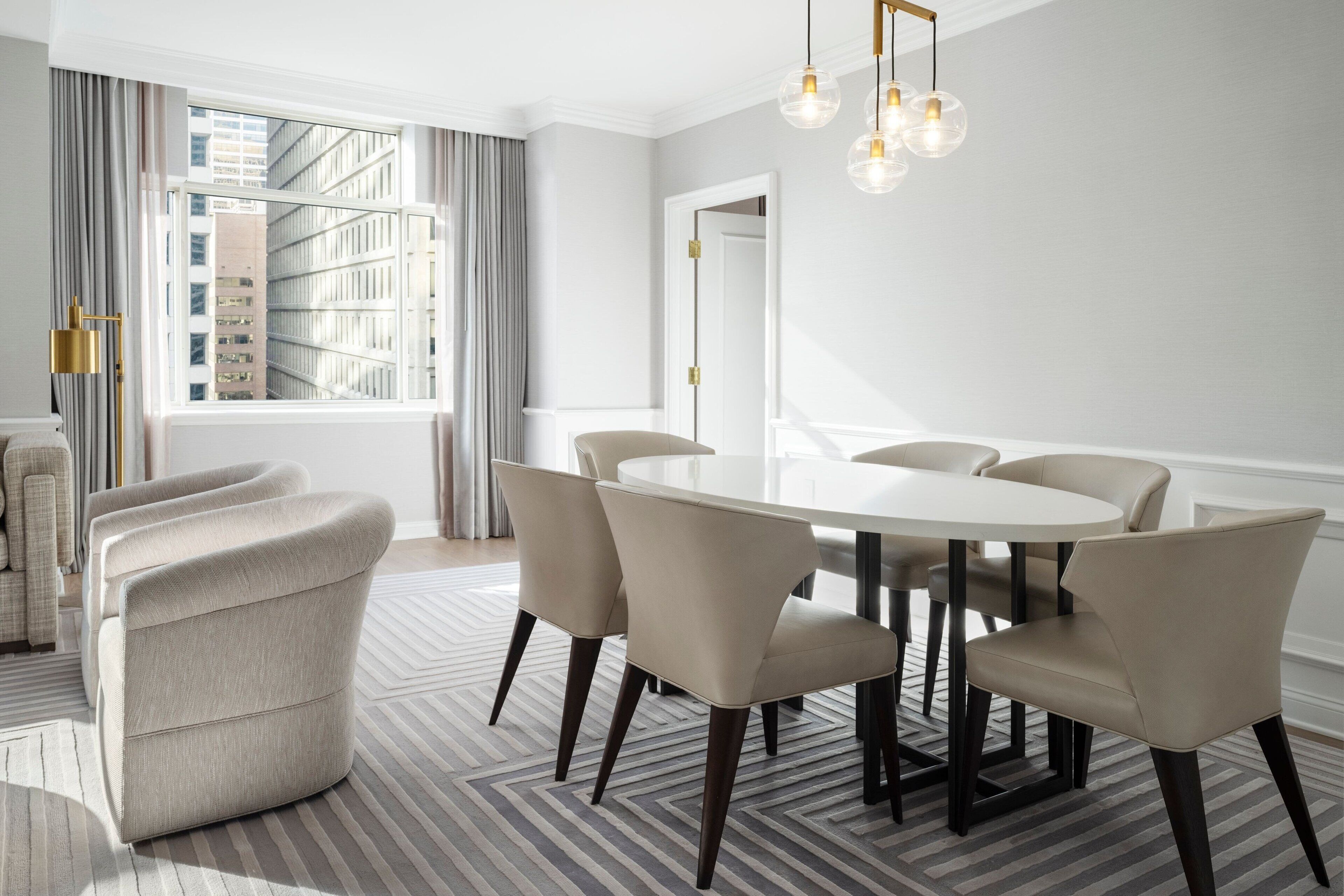 A modern dining area with a round table, six chairs, and a view of tall buildings through large windows.