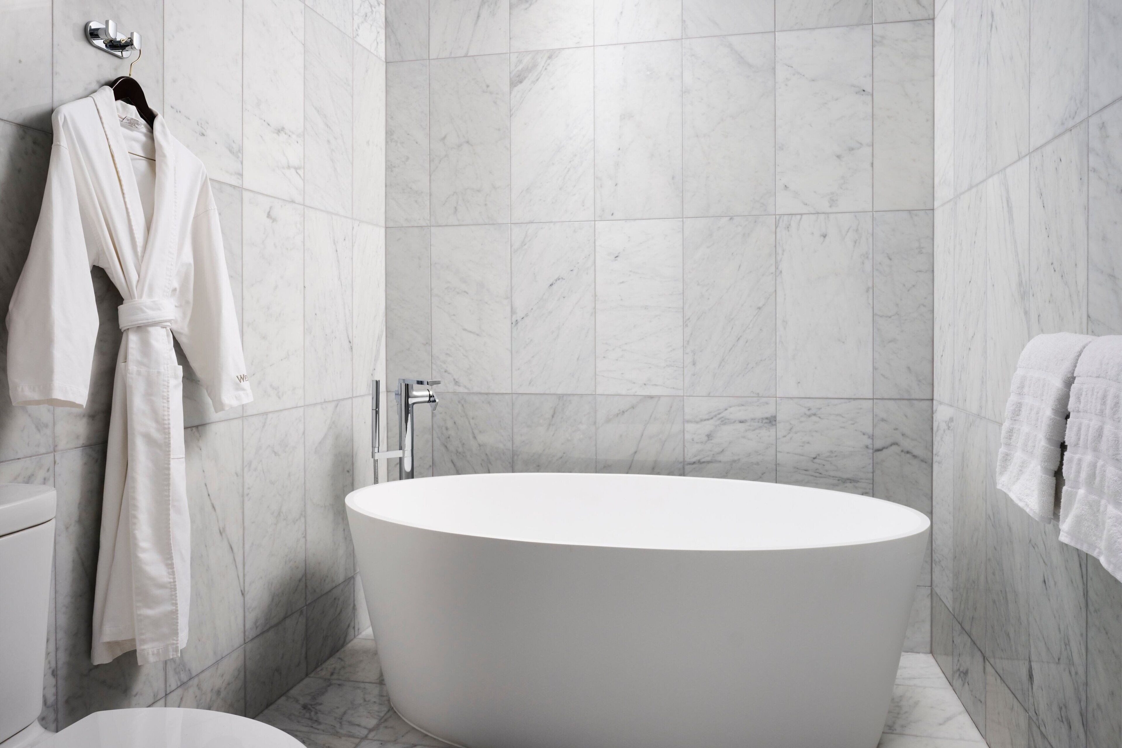A modern bathroom with a freestanding bathtub, marble walls, and a white robe hanging on a rack.
