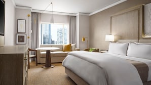 Ein Hotelzimmer mit einem großen Bett, einem Schreibtisch, einem Stuhl, einem kleinen Tisch und Blick auf die Stadtlandschaft.