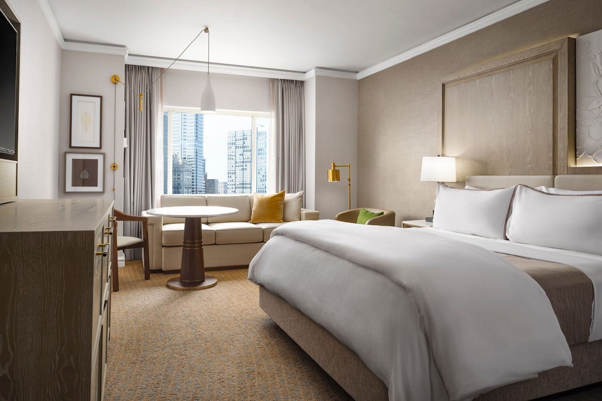 A hotel room with a large bed, a desk, a chair, a small table, and a view of the cityscape.