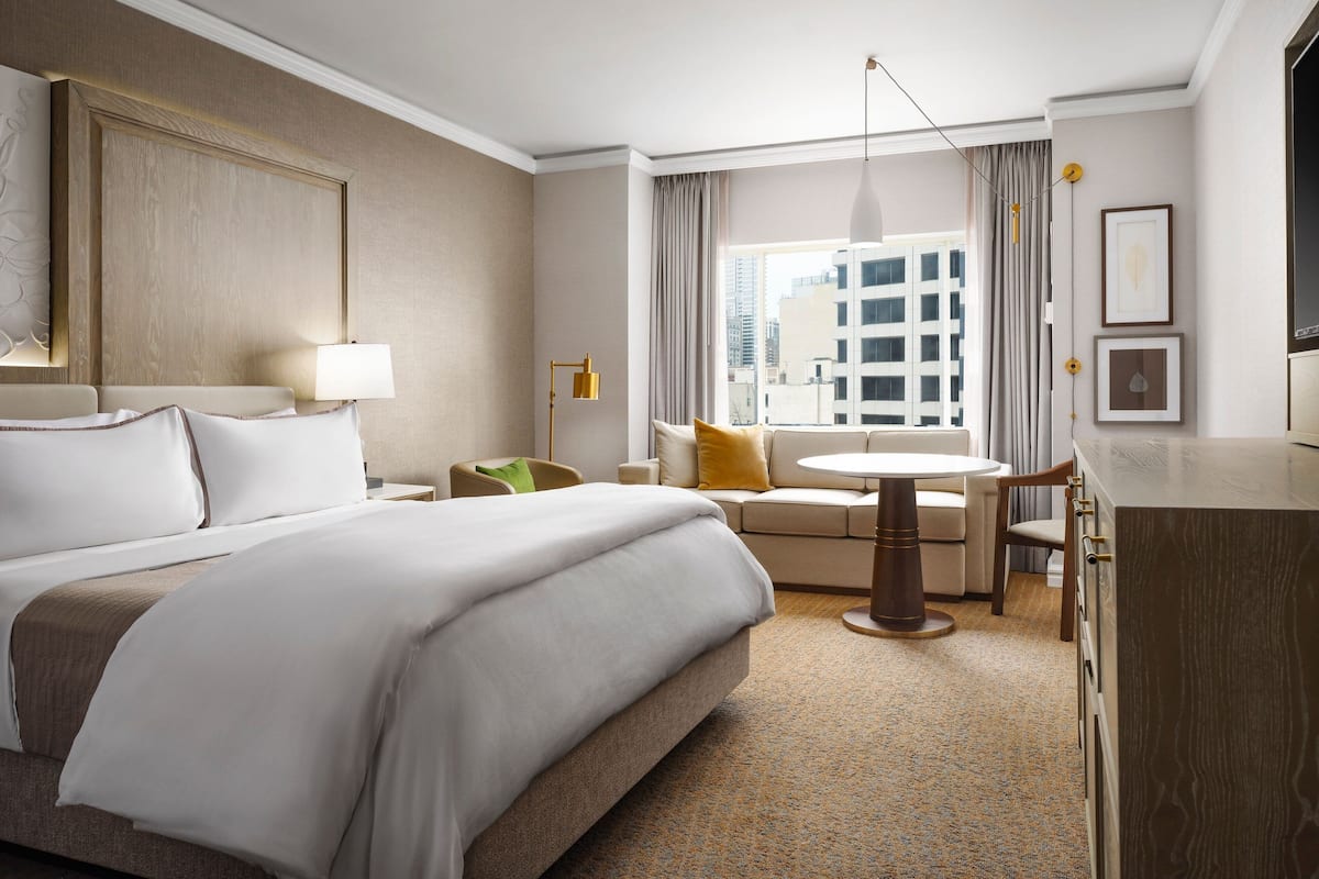 A hotel room with a large bed, a sofa, a dining table, and a view of the city through the window.