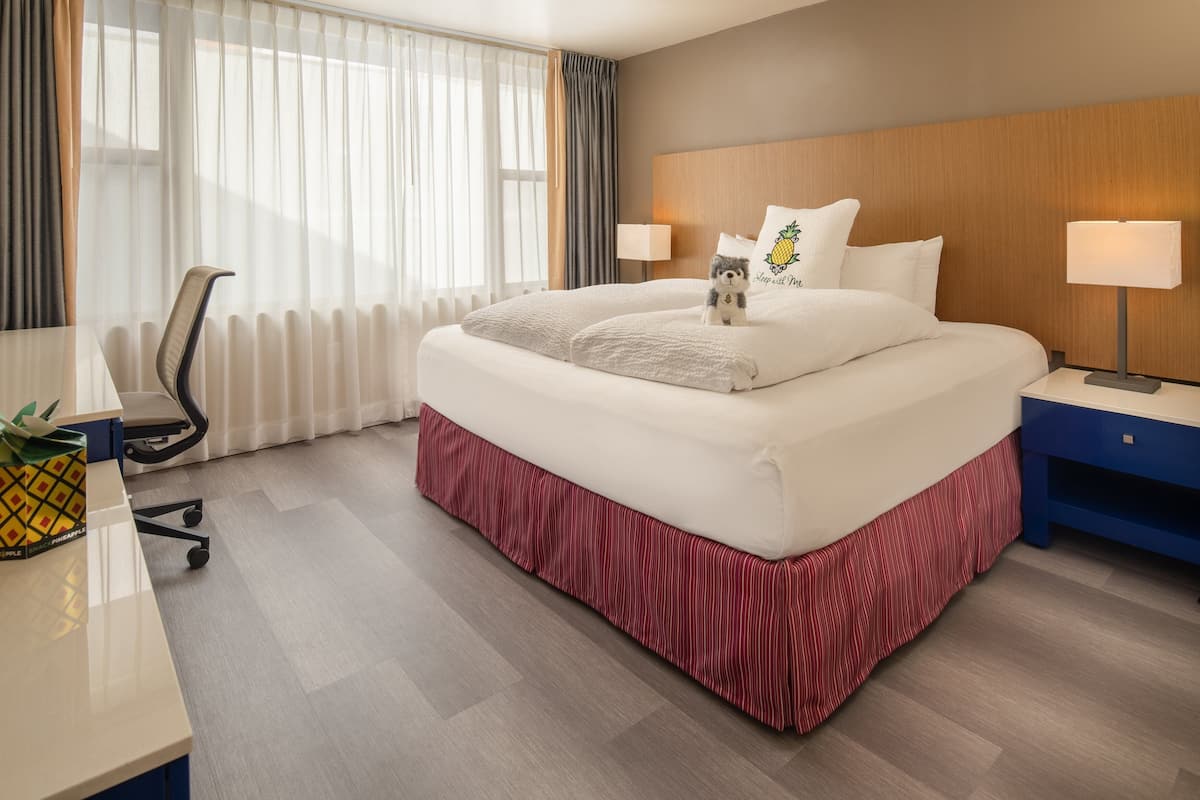 A hotel room with a bed, two bedside lamps, a desk with a chair, and a window with curtains.