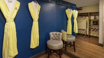 A hotel room with a blue wall, a mirror, and three yellow robes hanging on hooks. There is a grey chair and a small table in the room.