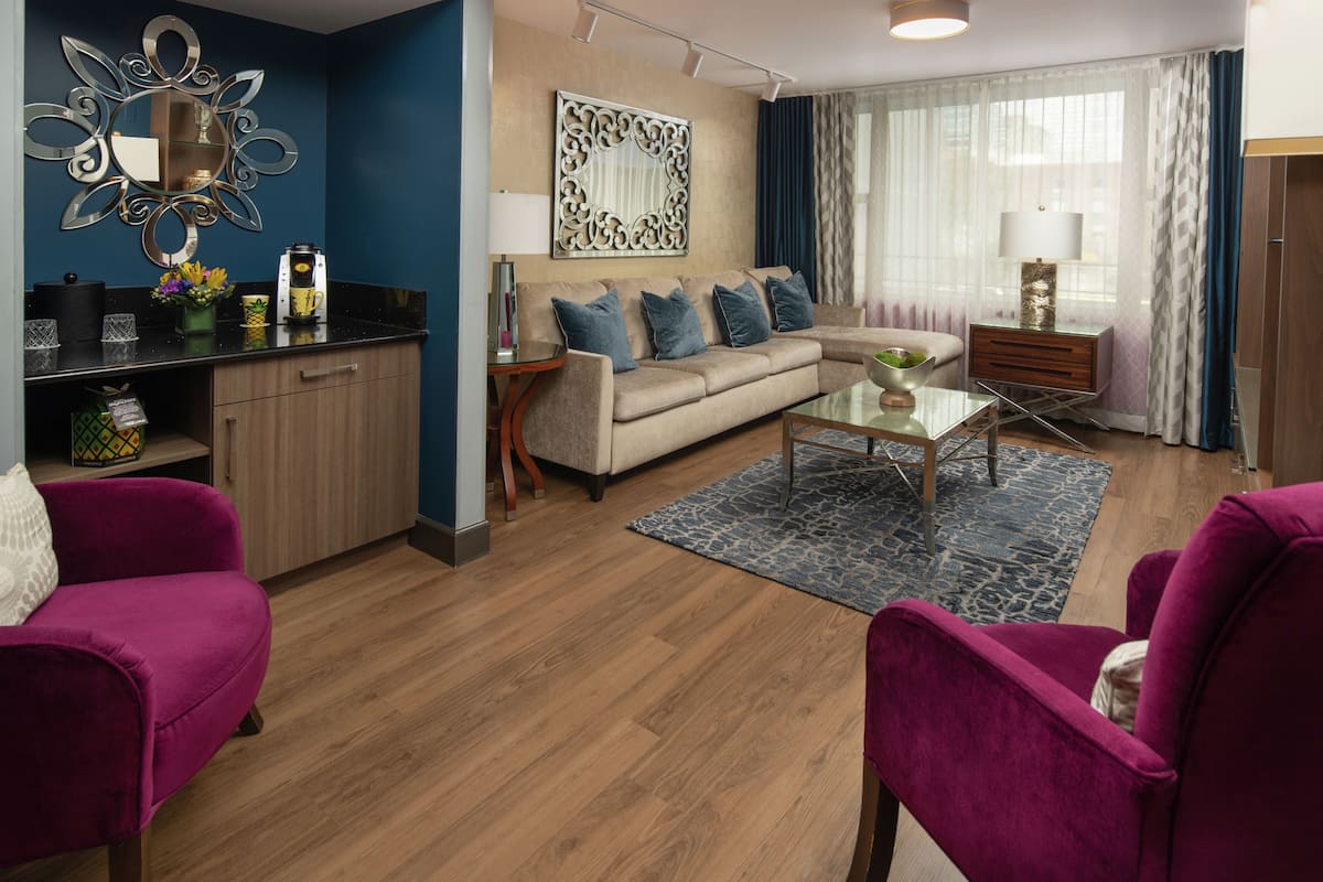 A modern living room with a beige sofa, a glass coffee table, and a purple armchair. There is a kitchen area with a black countertop and a wooden cabinet.