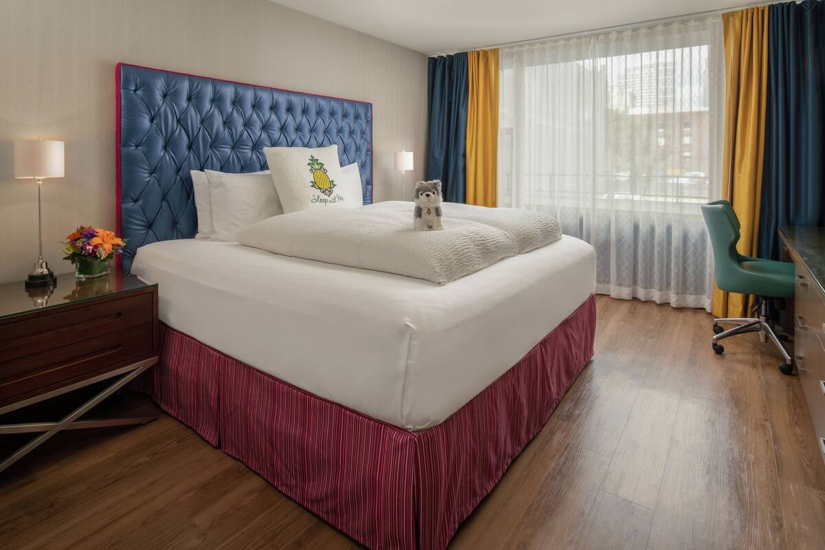 A hotel room with a tufted headboard, a bed with white bedding, a small desk with a lamp, and a view through sheer curtains.