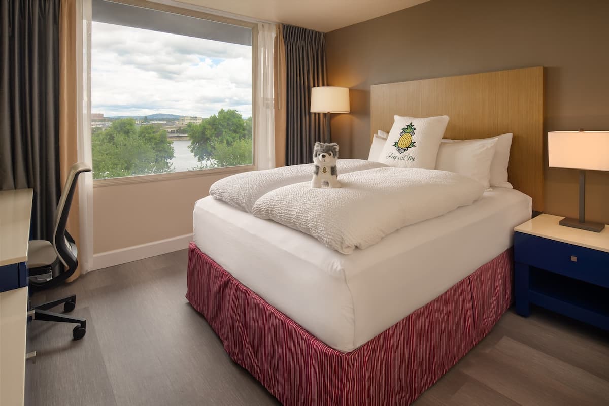 A hotel room with a large window, a bed with white bedding, a small desk with a chair, and a nightstand with a lamp.