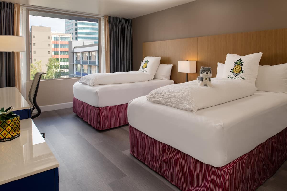 A hotel room with two beds, a desk, a chair, a lamp, and a window with a view of buildings.