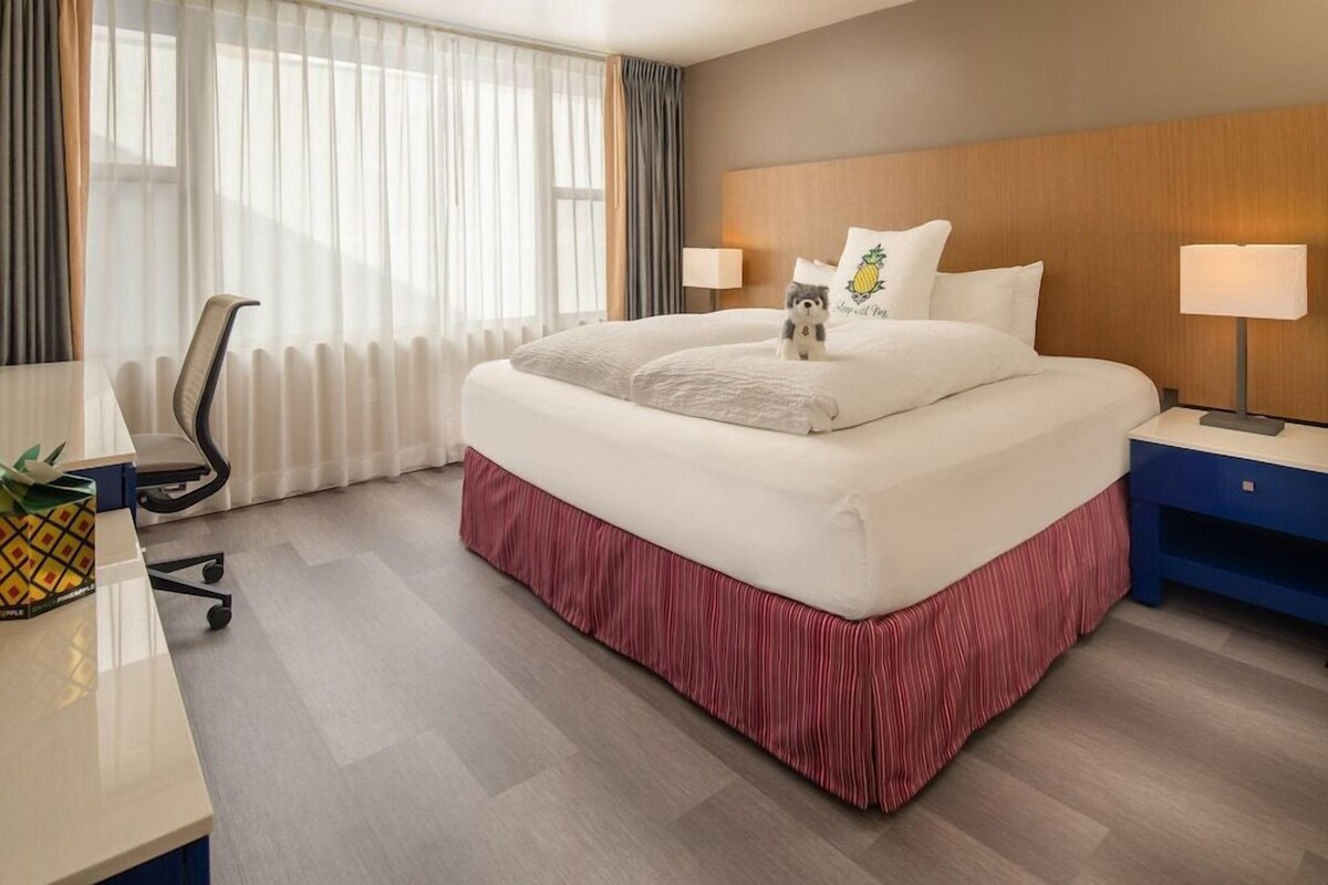 A hotel room with a bed, two bedside lamps, a desk with a chair, a small table with a pineapple, and a window with curtains.