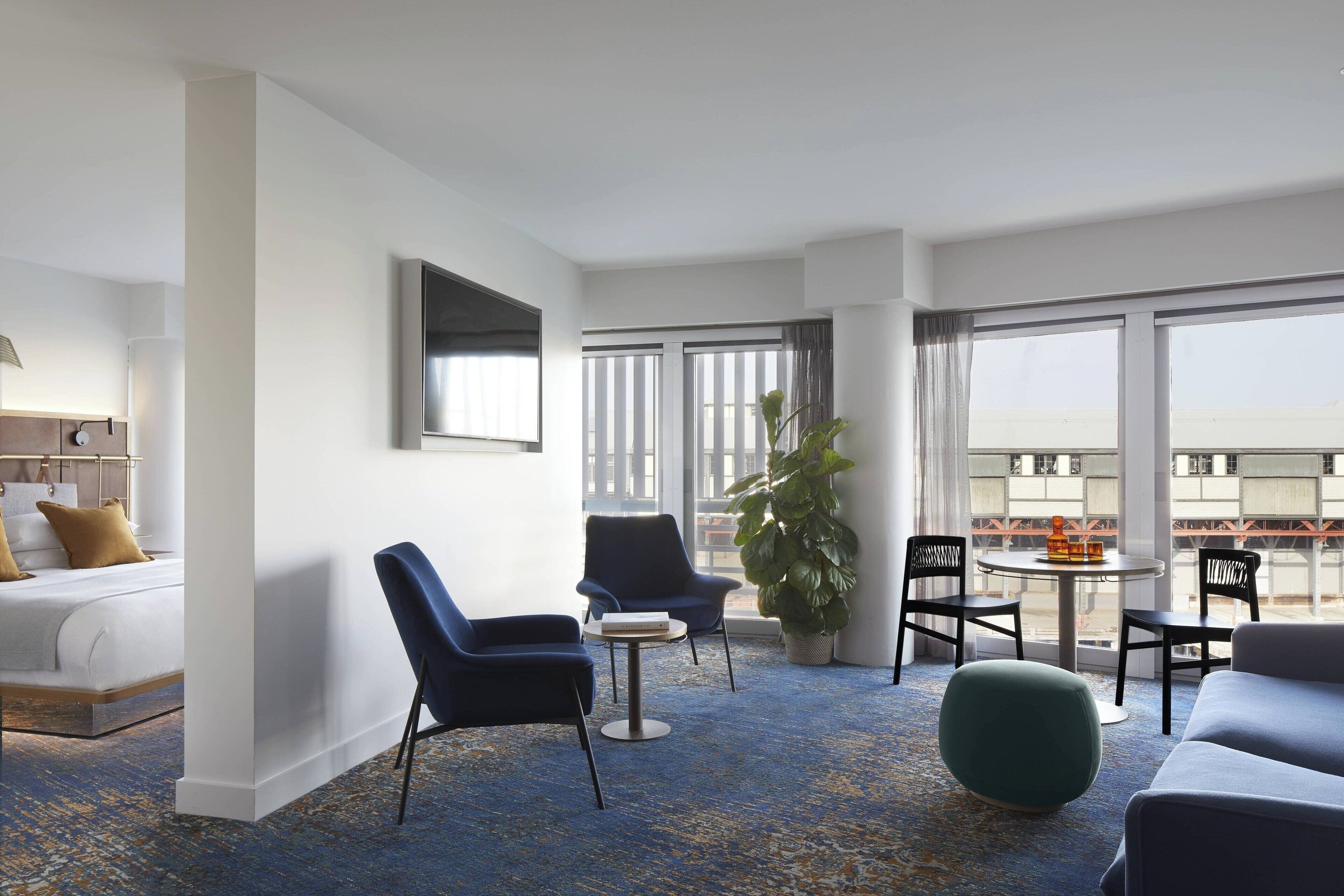 Ein Hotelzimmer mit Bett, einer Sitzecke mit Stühlen, einem kleinen Tisch und einem großen Fenster mit Blick auf die Stadt.