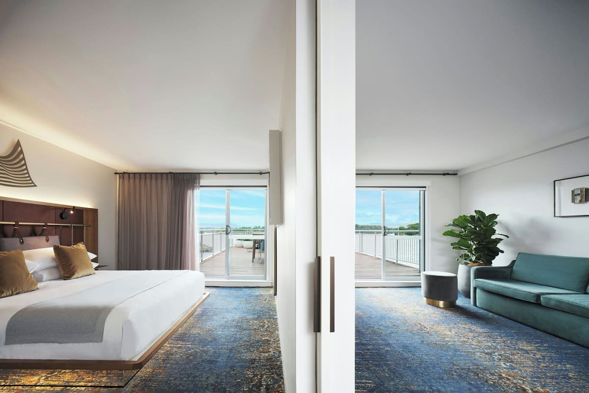 A hotel room with a large bed, a sofa, a potted plant, and a balcony with a view.
