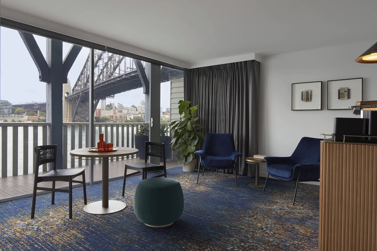 A modern hotel room with a large window overlooking a cityscape and a bridge. It includes a round table, two chairs, a sofa, and a potted plant.