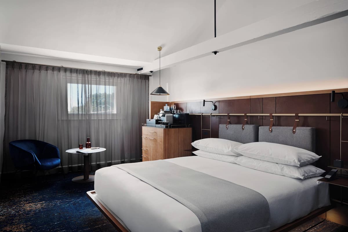 A hotel room with a large bed, a small table, a blue chair, a wardrobe, and a window with curtains.