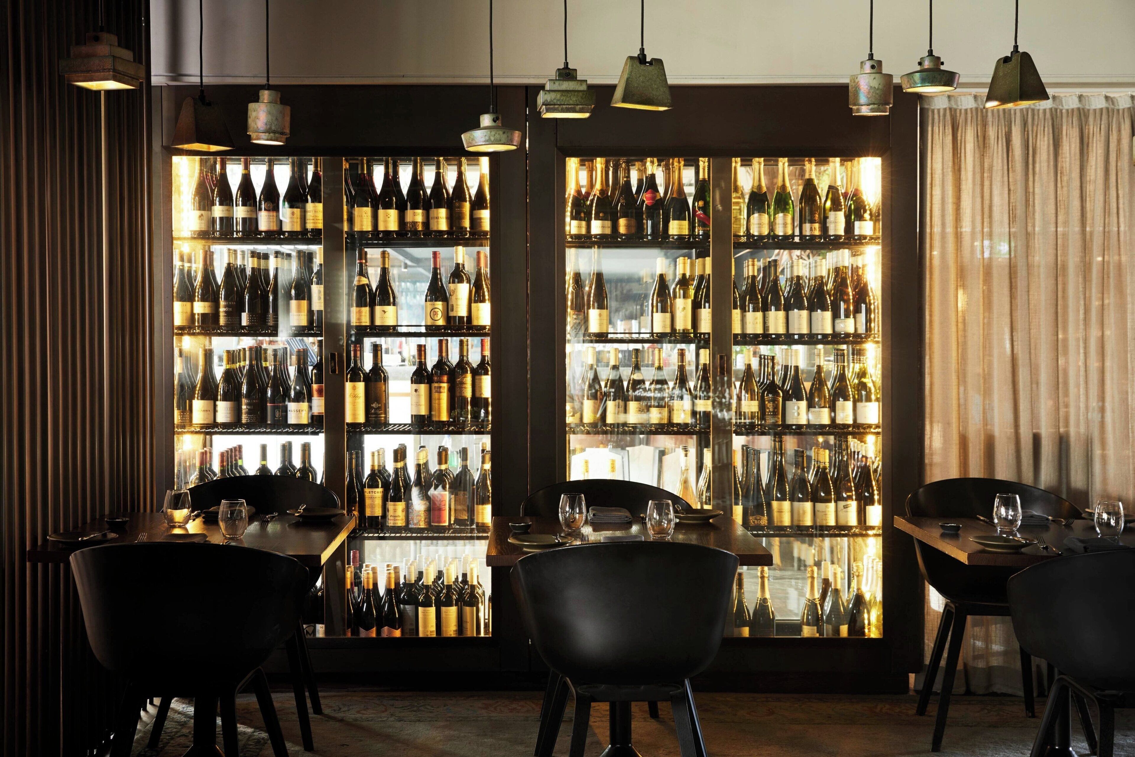 Una zona de bar con múltiples estantes de botellas de vino, mesas con sillas y lámparas colgantes.