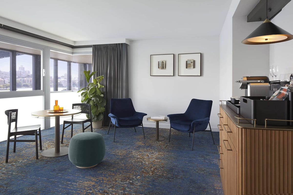 A modern hotel room with a large window, a round dining table with chairs, two blue armchairs, a small round table, a green ottoman, a wooden cabinet with drawers, and a hanging light fixture.