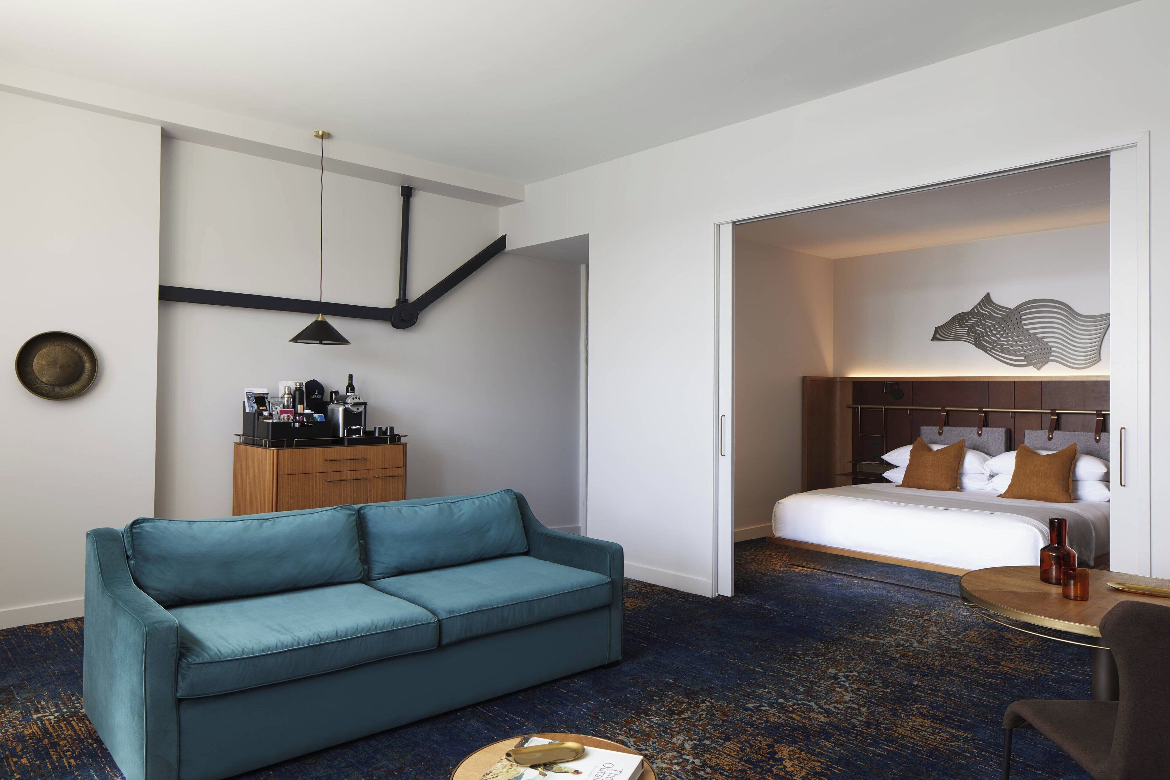 A hotel room with a teal sofa, a bed with brown pillows, a wooden cabinet with toiletries, and a wall-mounted artwork.