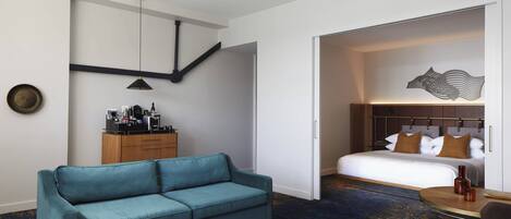 A hotel room with a teal sofa, a bed with brown pillows, a wooden cabinet with toiletries, and a wall-mounted artwork.