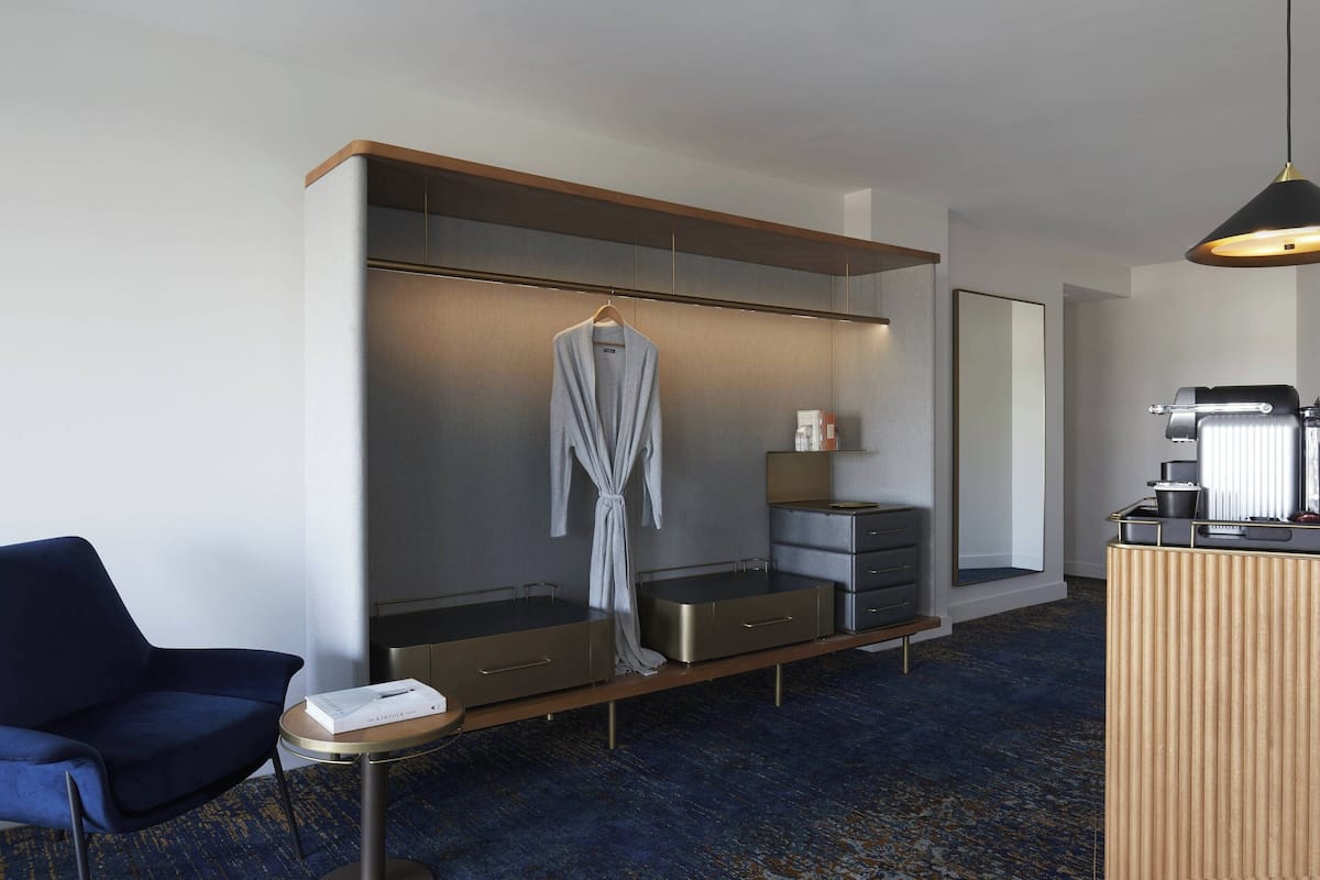 A modern hotel room with a large wardrobe, a seating area with a chair, a small round table, and a coffee machine.