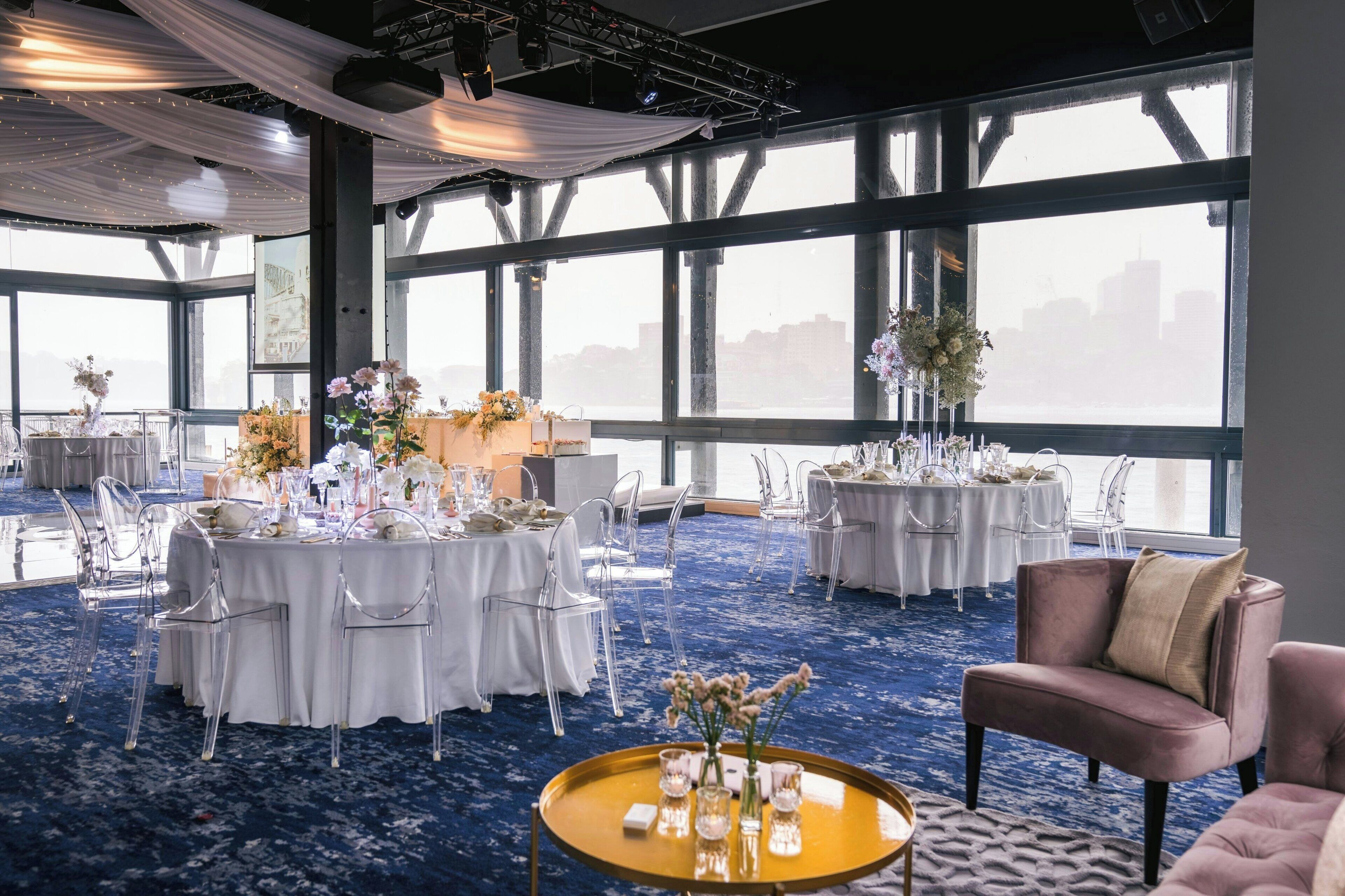 A spacious event room with round tables set for a formal event, large windows with a city view, and a mix of clear chairs and plush seating.