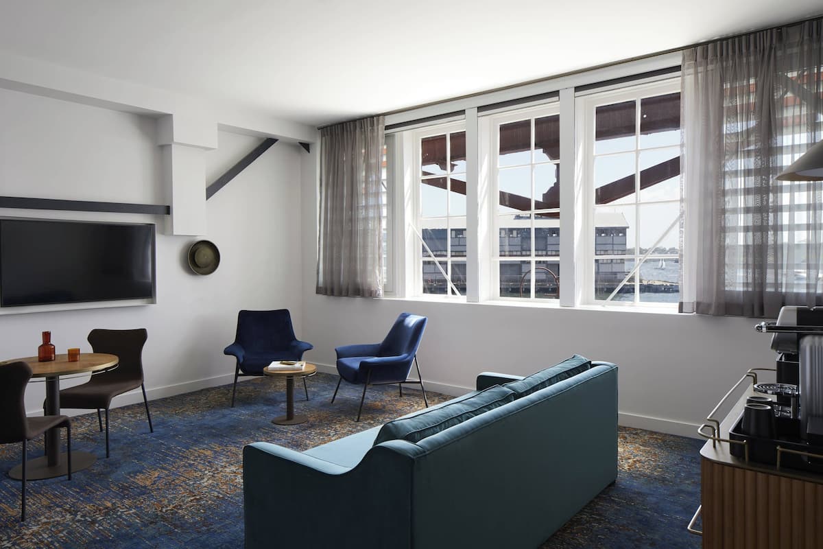 A modern hotel room with a large window, a sofa, two armchairs, a small table, and a flat-screen TV mounted on the wall.