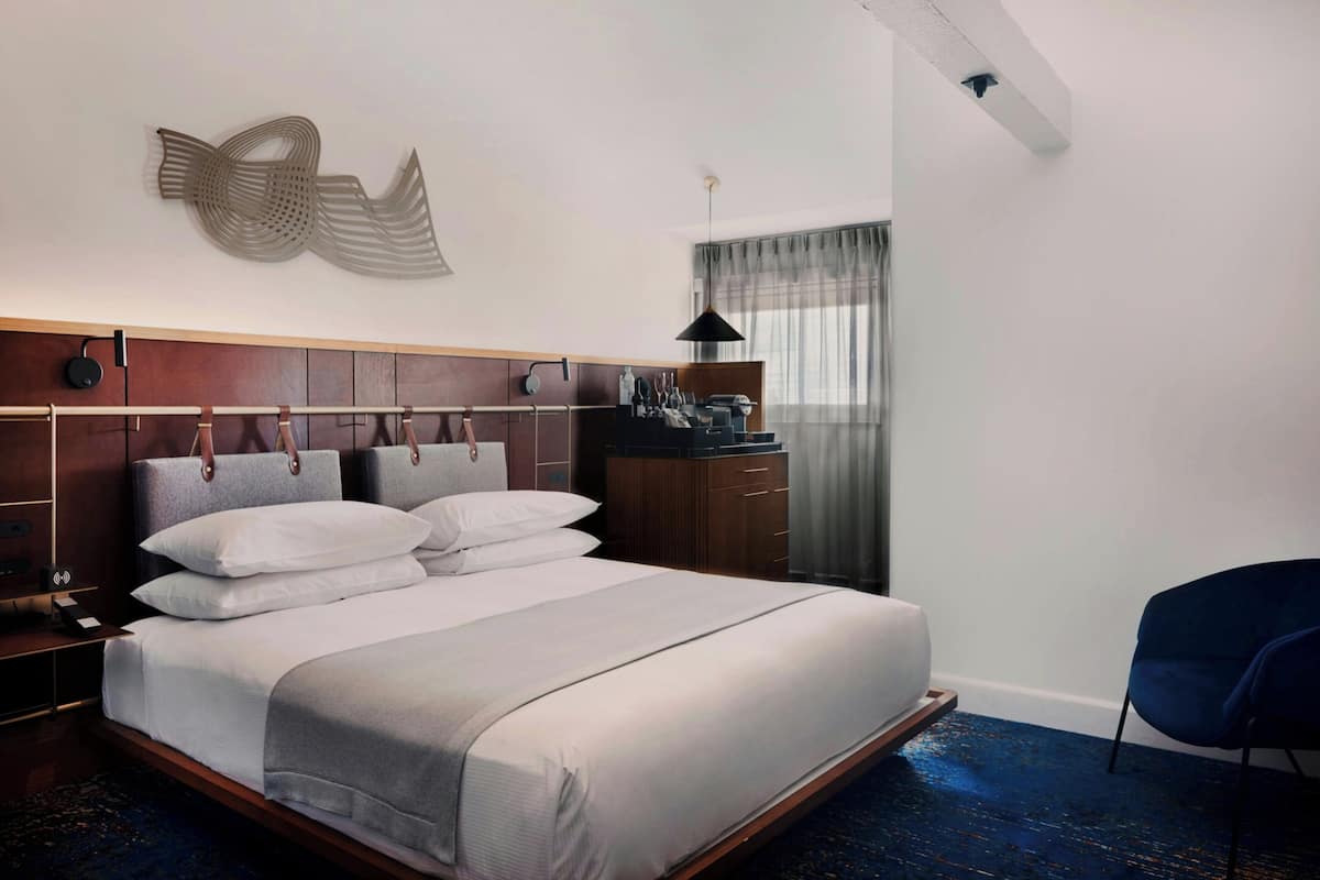 A modern hotel room with a large bed, a wooden headboard, a desk with a lamp, and a chair.