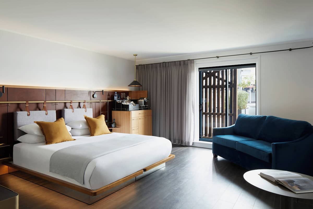 A hotel room with a large bed, a sofa, a small table, and a view of the outdoors through a sliding door.