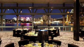 Un restaurante con mesas dispuestas para comer, vista a un horizonte urbano y un cuerpo de agua al fondo.