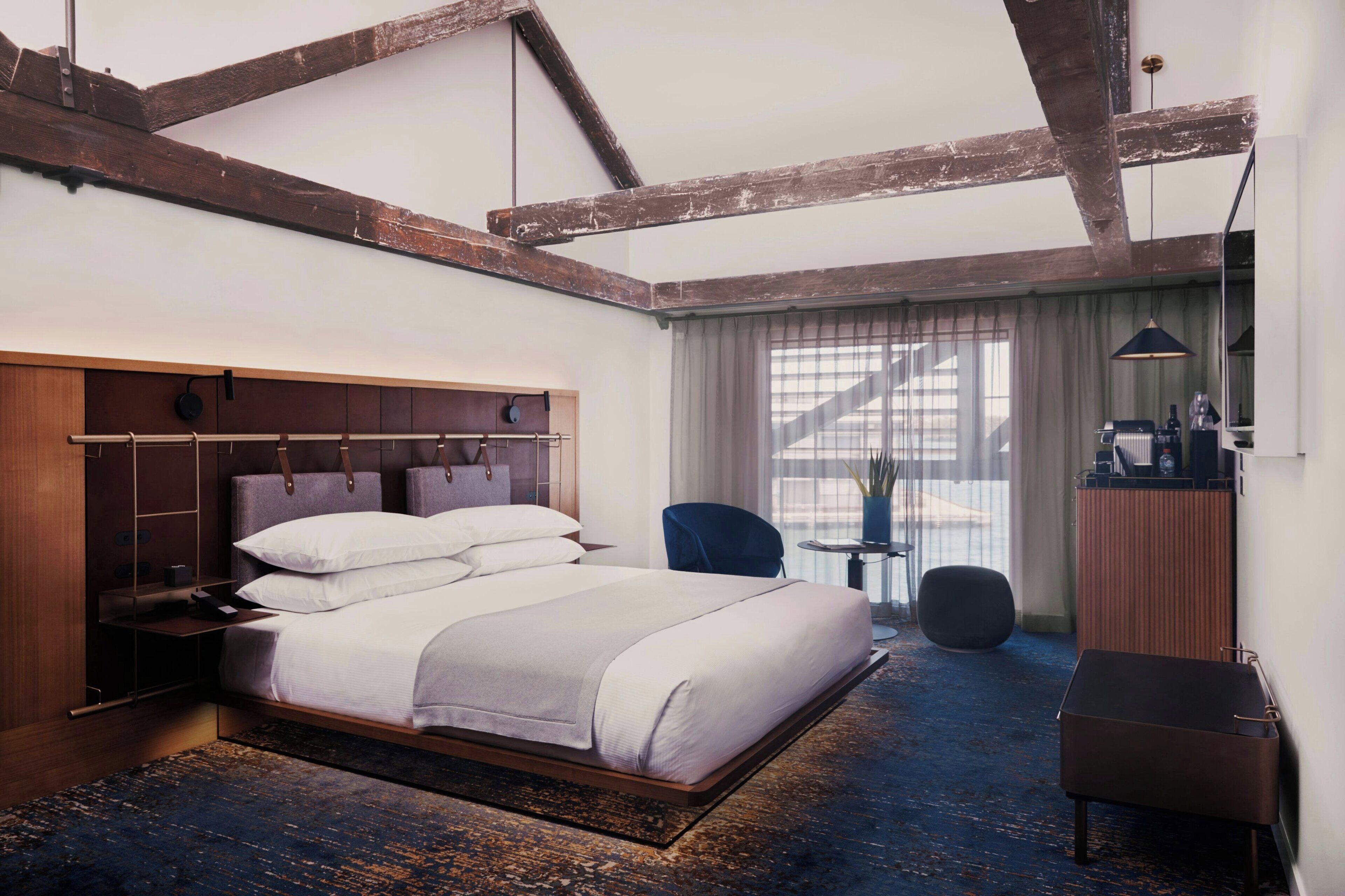 A modern hotel room with a large bed, wooden beams on the ceiling, a desk, and a chair.