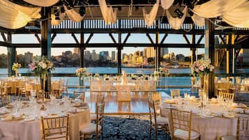 Un salón de banquetes con mesas redondas dispuestas para un evento formal, con vista a un paisaje urbano frente al mar.