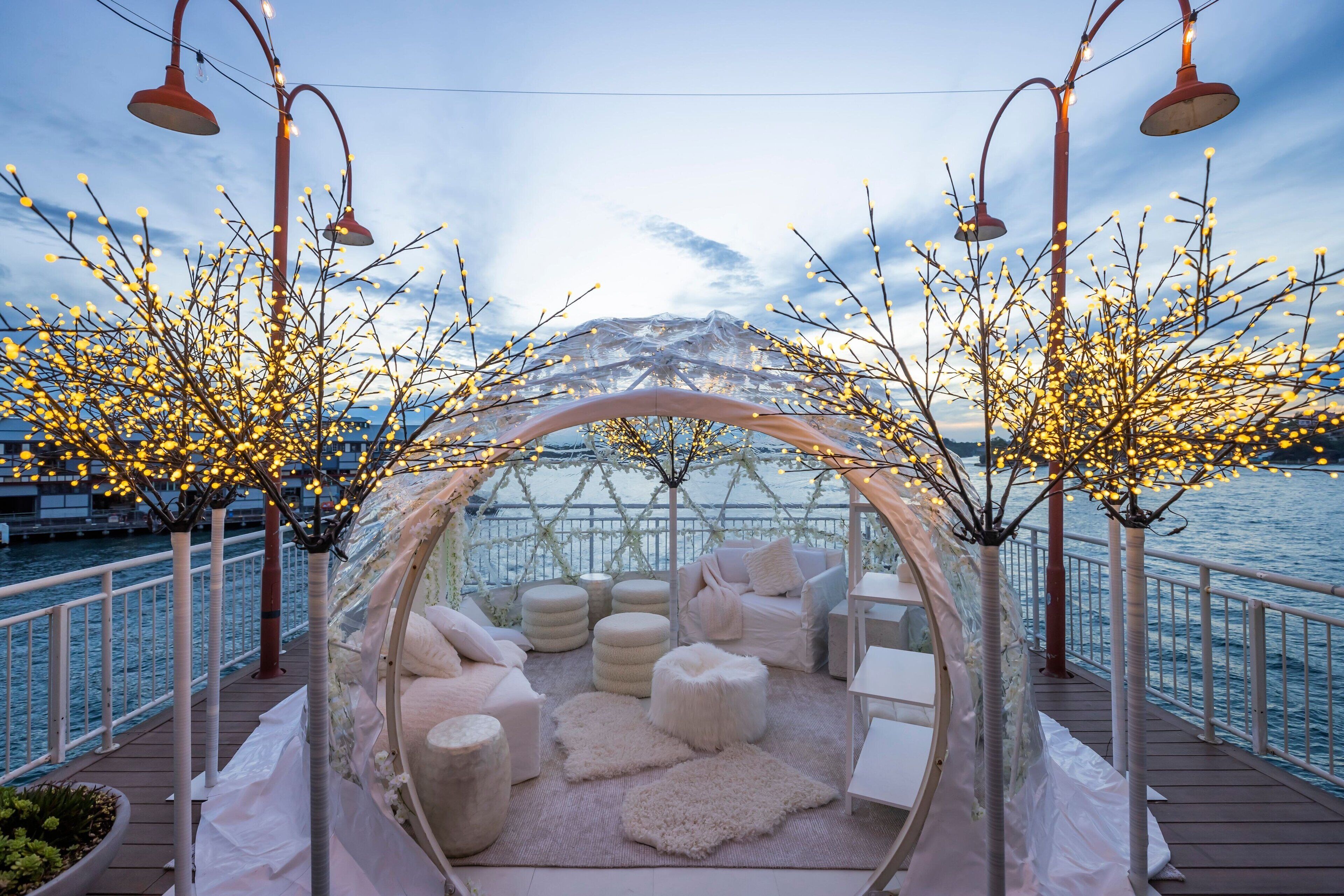 A covered outdoor seating area with a transparent canopy, cushioned seating, and a view of the water.