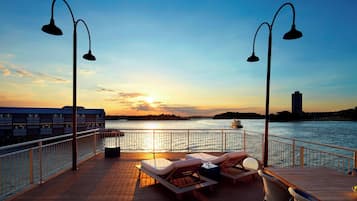 A wooden deck with lounge chairs and a table, overlooking a body of water with a sunset in the background.