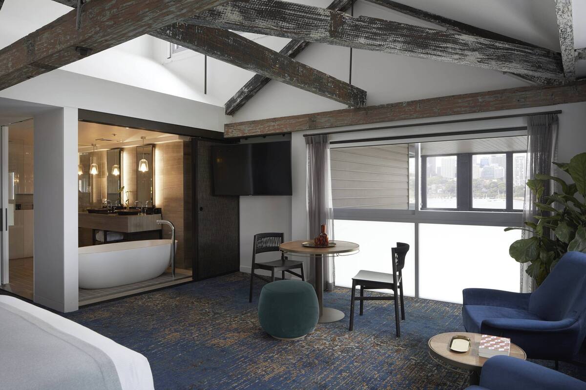 A modern hotel room with exposed wooden beams, a large window, a bathtub, a dining area with a table and chairs, and a large plant.