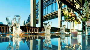 Una mesa con copas de cristal, una vela y un jarrón con flores, reflejando un edificio moderno con amplias ventanas y una estructura metálica al fondo.
