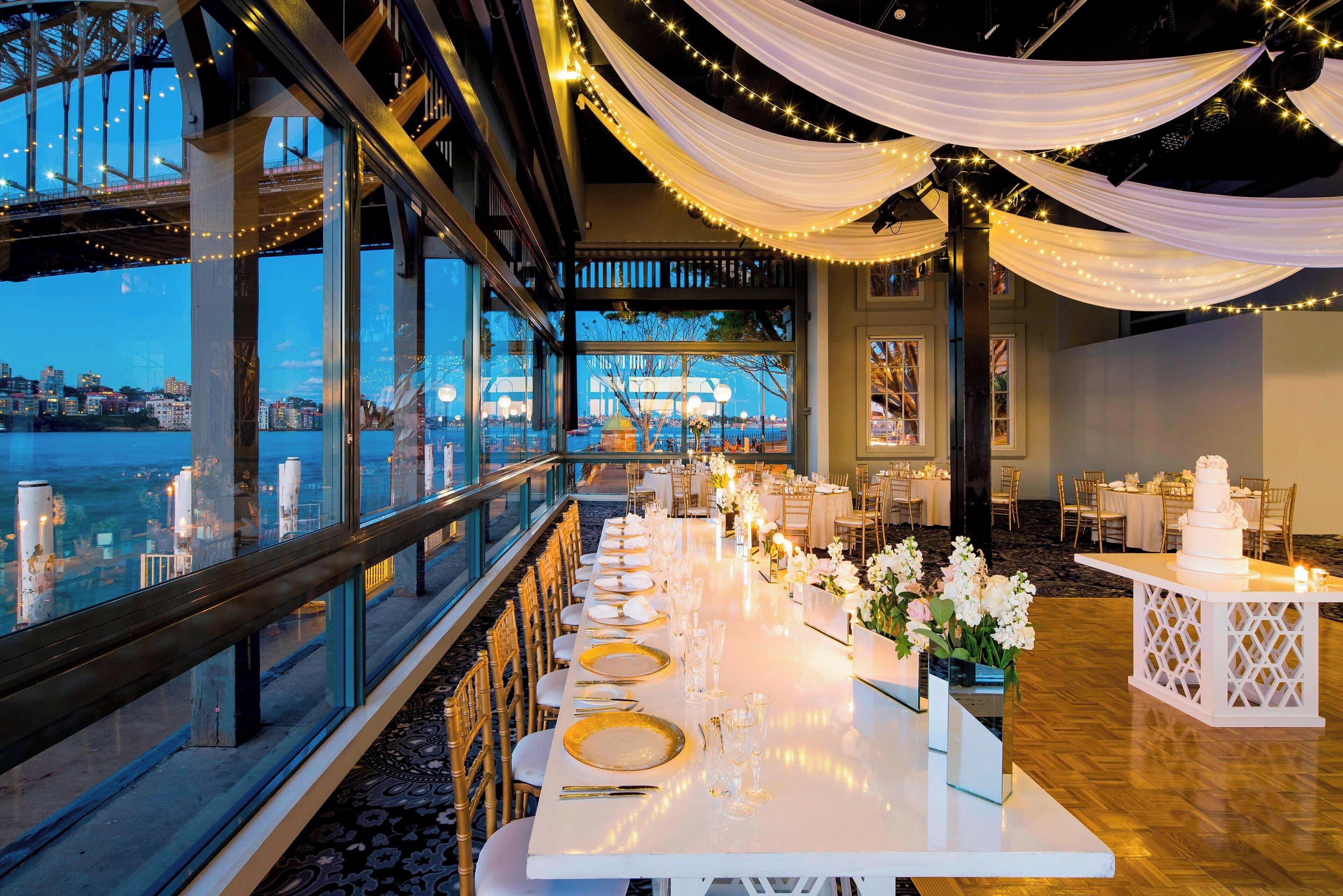 Une longue table à manger, dressée pour un événement formel, avec vue sur la ville, recouverte d’un tissu blanc et illuminée de guirlandes lumineuses.