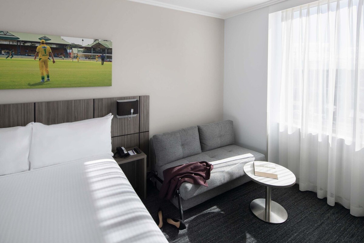 A hotel room with a bed, a grey sofa, a small round table, a TV, and a window with sheer curtains.