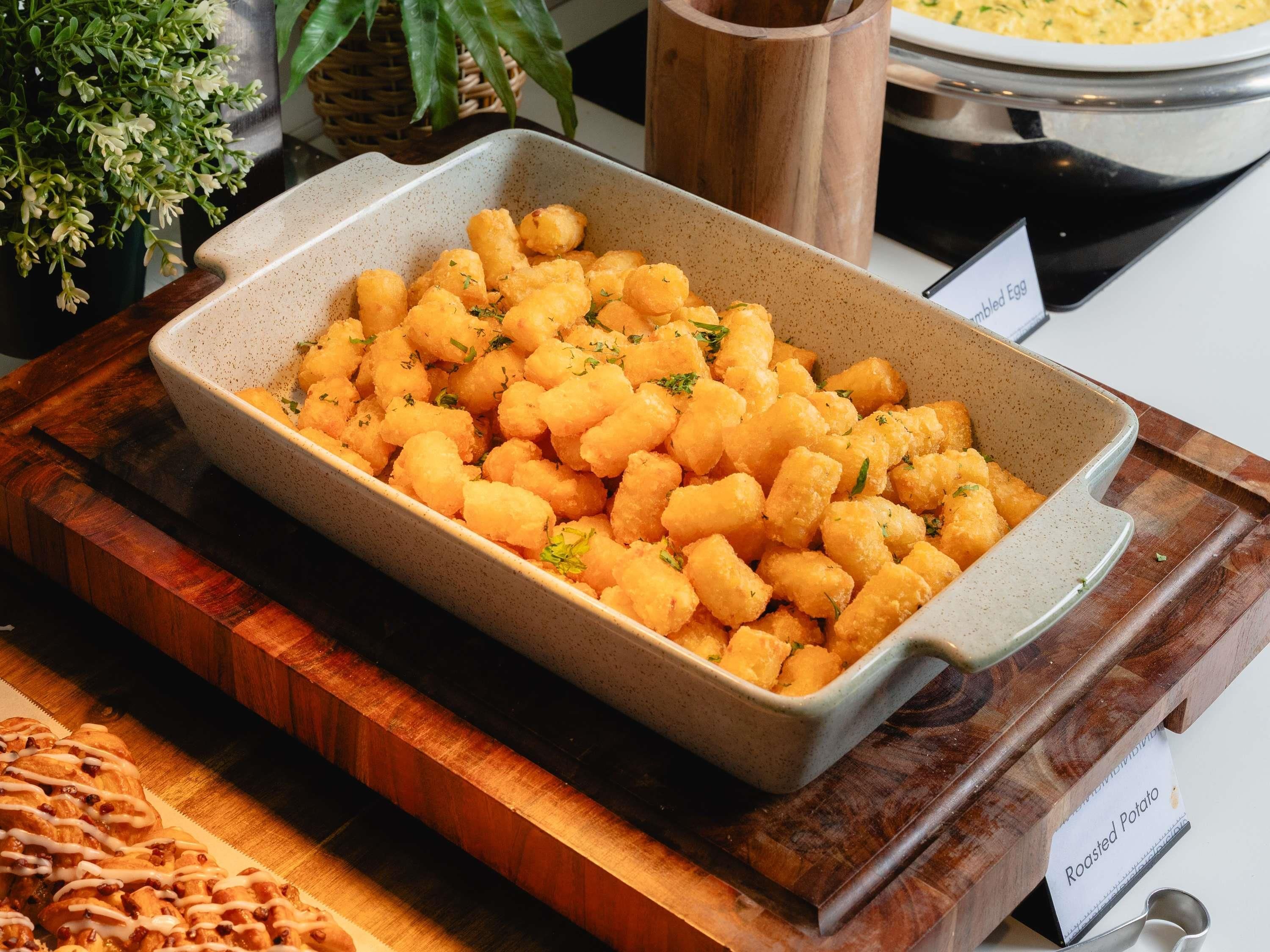 A dish of tater tots garnished with herbs, placed on a wooden board with a label indicating 'Roasted Potato'.