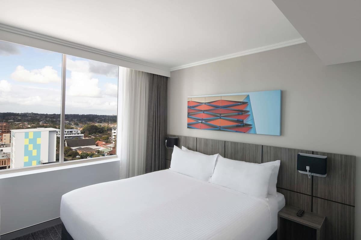 A hotel room with a large bed, a nightstand, a wall-mounted lamp, and a window with a city view.
