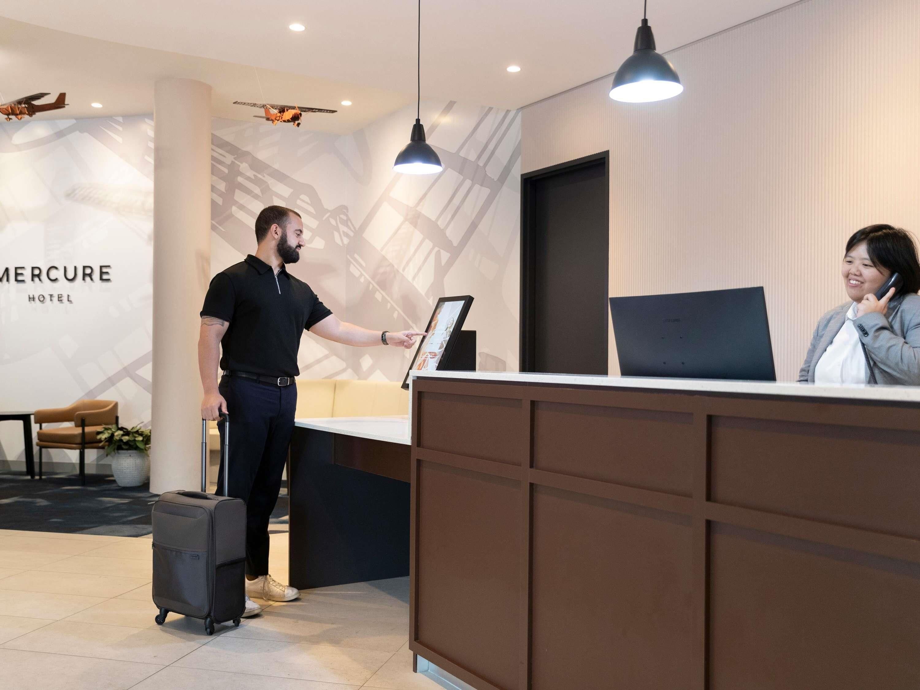 A hotel reception desk with a digital display, a seated receptionist, and a standing guest with a suitcase.